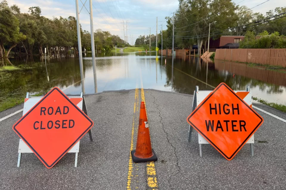 Orange and black "Road Closed" and "High Water" signs blocking a flooded roadway in Florida after a hurricane.